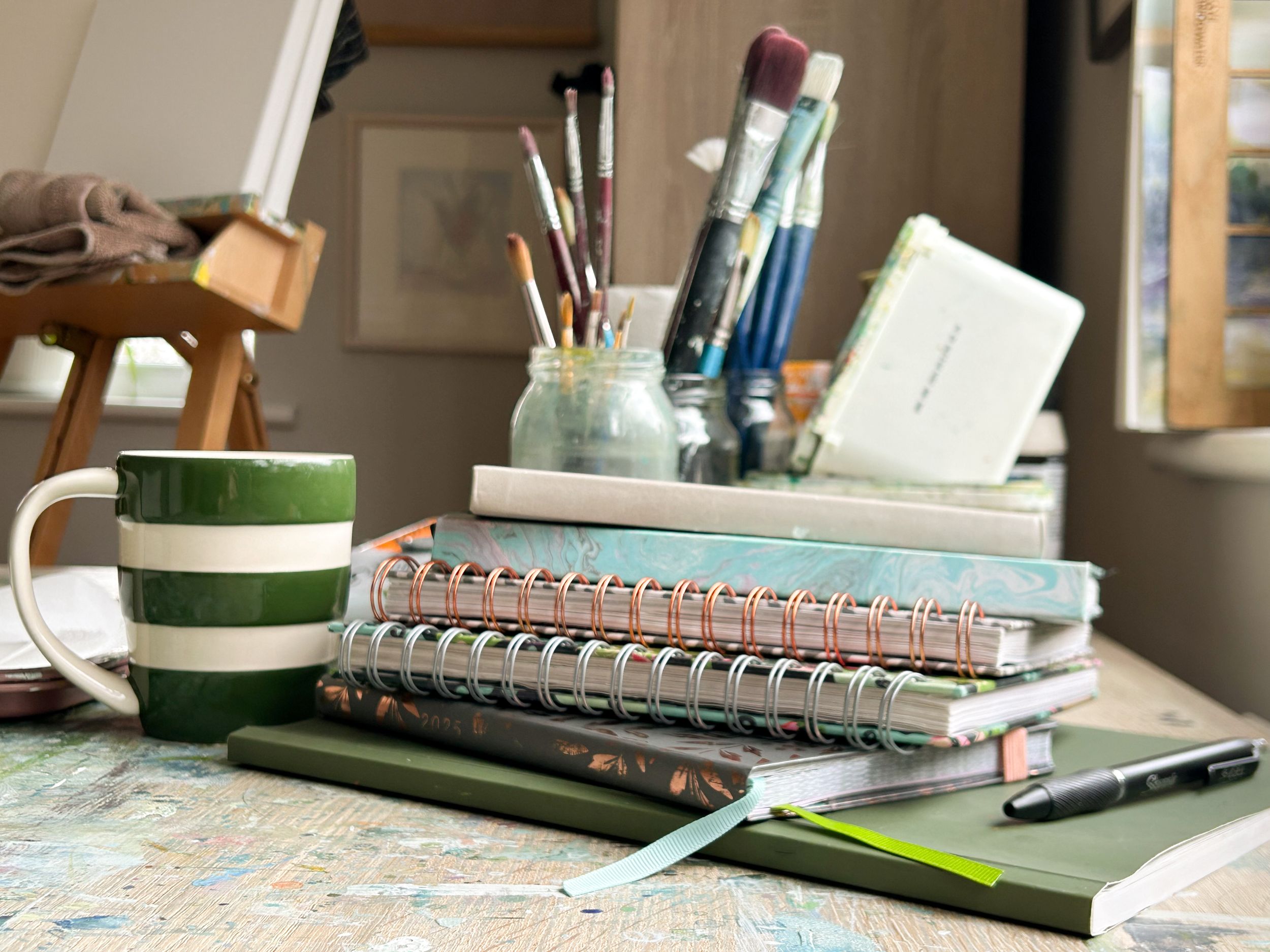 studio scene of painting table piled with notebook journals mug of tea and painting stuff